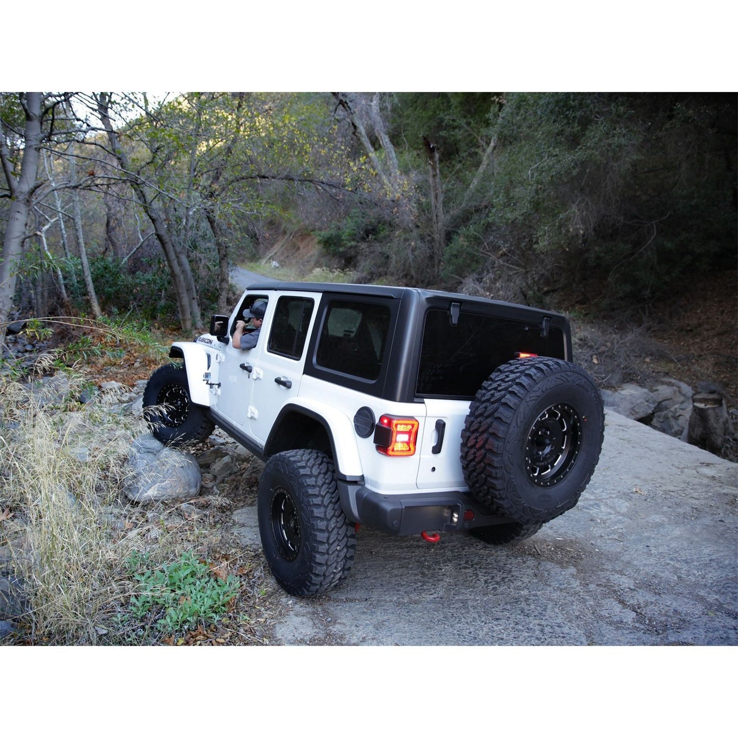SMITTYBILT | JL Tire Relocation Bracket 2018-Up Jeep Wrangler JL Smittybilt |7721 displayed on a white Jeep navigating rocky terrain.