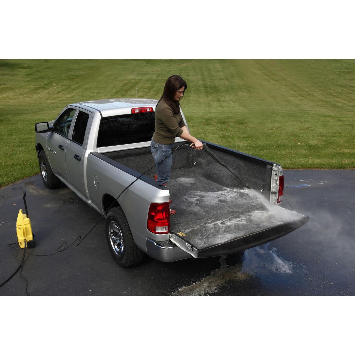 A woman cleaning the truck bed of a silver Ram 1500 with a pressure washer, showcasing the BedRug 02-18 - Charcoal (19-23 Classic) Ram 1500/03-24 2500/3500 8'.